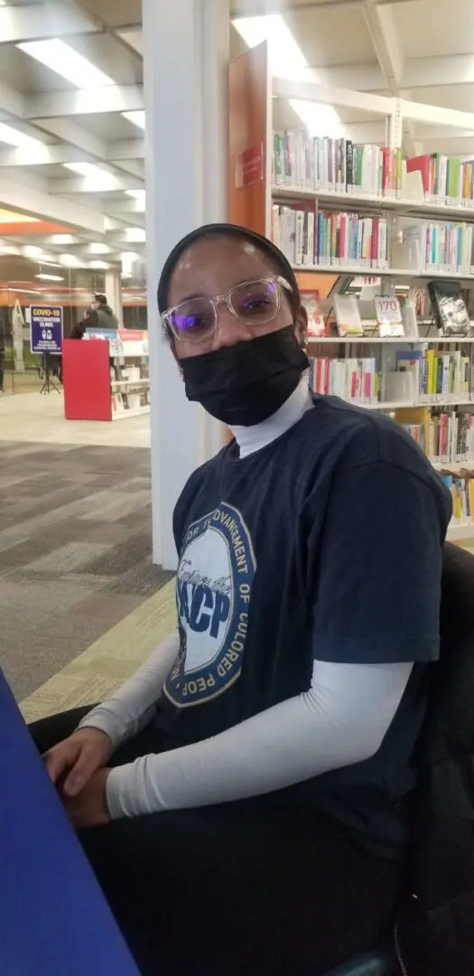 A person wearing glasses and a mask sitting in front of books.